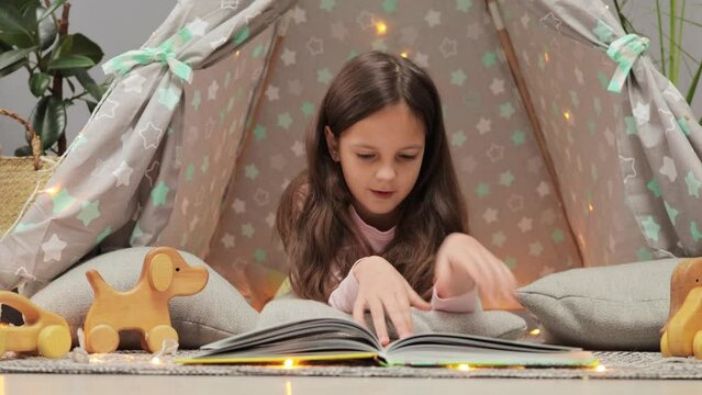 Surprised amazed little girl with dark hair reading book with astonished expressing being impressed looking at pictures with interest lying in children's teepee shelter at home.