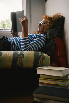 Side view of elderly woman reading book while lying on bed at home