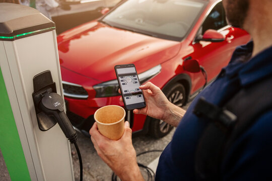 Man holding disposable cup using smart phone at electric car charging station