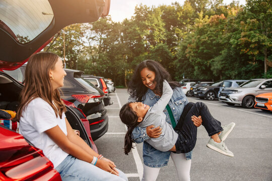 Girl sitting in trunk looking at mother having fun with daughter near car