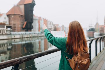 Attractive young female tourist is exploring new city. Redhead girl with backpack holding a paper...
