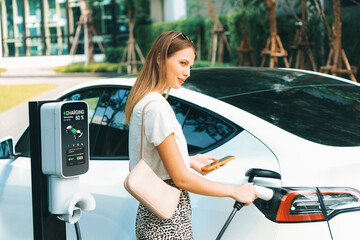 Young woman travel with EV electric car charging in green sustainable city outdoor garden in...