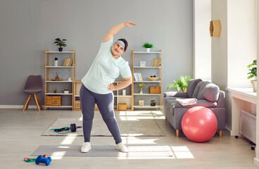 Full length photo of a happy funny young smiling fat overweight woman wearing sportswear doing fit exercise in the living room at home. Workout sport, fitness and body positive concept.