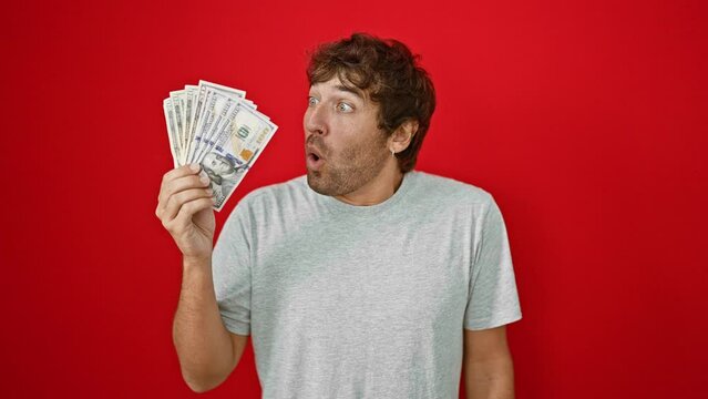 Shocked young man holding us banknotes, agape in amazement, fear, and disbelief against a stark red backdrop! wow, what a surprised face!