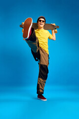 Full length portrait of cool dude rastaman, skateboarder holding skateboard and posing against blue...