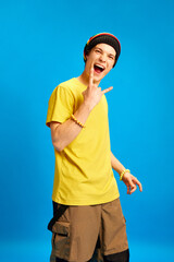 Happy shouting young man wearing rastaman, reggae style outfit showing rock sign against blue...