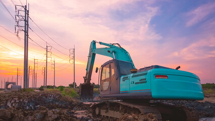 Excavator is leveling the ground in large yard for construction area of industrial building with row of electric poles against sunset sky in evening time