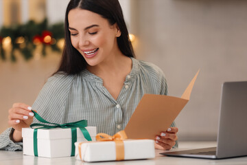 Naklejka premium Celebrating Christmas online with exchanged by mail presents. Smiling woman opening gift box during video call on laptop at home