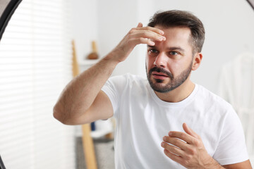 Confused man with skin problem looking at mirror indoors