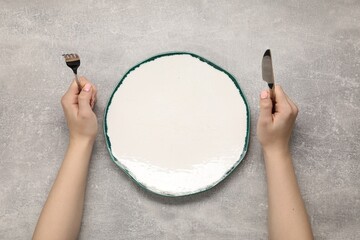 Woman holding fork and knife near empty plate at grey textured table, top view