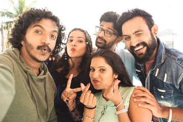 Group of young Indian friends taking selfie with grimacing faces by looking camera during reunion party - concept of friendship bonding, zen z lifestyle and social media sharing