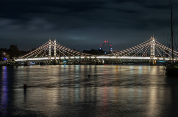Fototapeta premium Nocturnal view of floodlit Albert Bridge from Chelsea Bridge with the reflection of the light in very calm water of River Thames. Lit up at Night Illuminated Albert bridge in west London at night. Spa