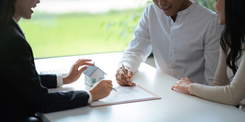 Young Asian couple making contract with house sale agency. man and his wife sitting signing the contract next to him looking the contract document with smile. real estate agreement successful concept
