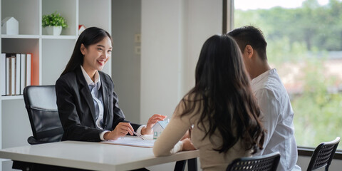 Young Asian couple making contract with house sale agency. man and his wife sitting signing the contract next to him looking the contract document with smile. real estate agreement successful concept
