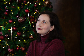 woman smiles thoughtfully against background of decorated Christmas tree....