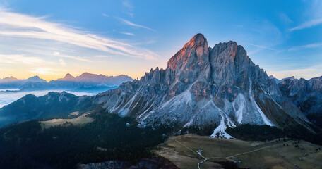 Sass de Puta mountain peak at Passo delle Erbe pass against the Dolomite peaks in the background, inverse cloud cover in the valley, blue sky, sunrise. Aerial drone view, large panorama. 