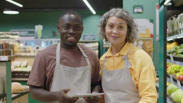 Medium portrait of two diverse male and female supermarket workers in aprons looking at digital tablet and having conversation then smiling at camera standing together in aisle with grocery