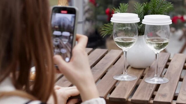 glass with sparkling wine and coffee cover on outdoor cafe table.girl taking photos,using smartphone,typing message.for christmas tree branches, happy new year.