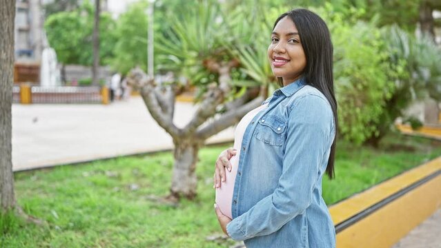 Joyful young latin woman, happily expecting motherhood, confidently touching her pregnant belly, smiling in sunlight, standing casually amidst city's green park