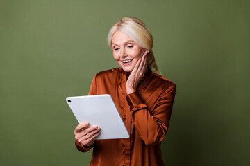 Photo portrait of attractive pensioner woman hold tablet shocked wear trendy brown satin clothes isolated on khaki color background
