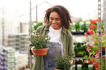 Smiling woman gardener holding flower pot while standing on greenhouse yard background - obrazy, fototapety, plakaty