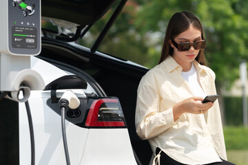 Young woman use smartphone to pay for electricity at public EV car charging station green city...