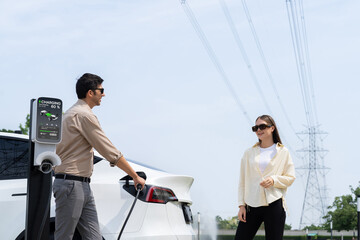 Young couple recharge EV car battery at charging station connected to power grid tower electrical...