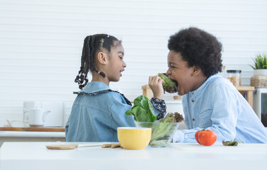 African little cute sister feeding smiling brother fresh green salad vegetable at home kitchen....