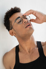 Fototapeta premium Portrait of relaxed and stylish young cuban man in black sleeveless t-shirt touching eyeglasses