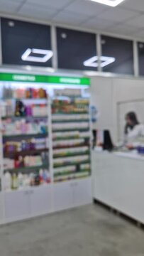 Blurred view inside of pharmacy with female pharmacist at workplace around stocked shelves with medicine. Healthcare and cosmetics industry defocused background