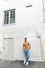 Full length of muscular and shirtless cuban man in blue jeans posing near building in Miami