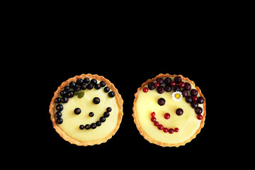 Smiling fruit cakes. Shortbread tartlets with vanilla custard cream, black and red currant and fruits of amelanchier. Isolated on black background, copy space for text. Creative baking