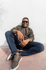 cheerful african american man in stylish sunglasses sitting near white wall