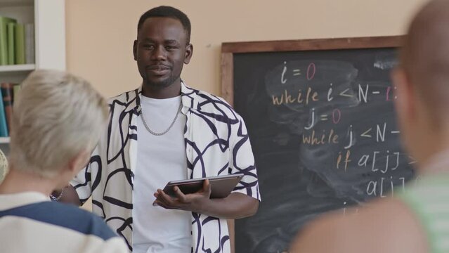 Young African American student holding digital tablet and talking to group mates while standing by chalkboard during programming class in college