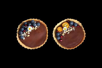 Shortbread tartlets with chocolate pudding, fresh blueberries, pomegranate seeds, kumquat slices, cashew nuts and edible daisy flowers. Isolated on black background