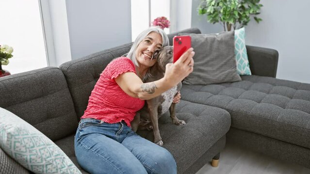 Joyful middle age woman enjoying her pet dog's company, sitting on sofa at home. smiling grey-haired lady having selfie fun with her smartphone.