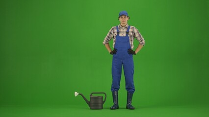 Portrait of farmer in working clothing on chroma key green screen. Gardener standing holding hands on waist, watering can next to him.