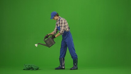 Portrait of farmer in working clothing on chroma key green screen. Gardener standing holding watering can and pouring water onto plants.