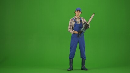 Portrait of farmer in working clothing on chroma key green screen. Gardener standing posing at the camera holding chainsaw.