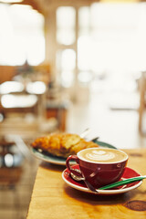Cappuccino with artful foam in a red cup on a wooden table, blurred bakery background