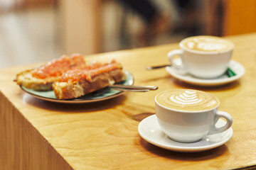 Latte with art on a wooden table, accompanied by a plate of bread with tomato