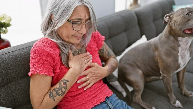 Heart-struck grey-haired middle age woman suffers attack in her cozy living room, sitting with treasured dog on comfortable sofa.