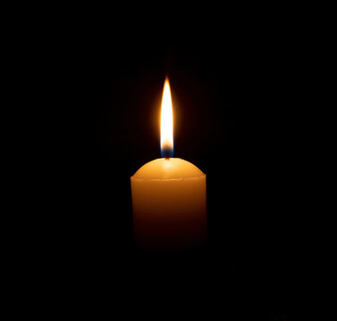 A single burning candle flame or light glowing on a big yellow candle on black or dark background on table in church for Christmas, funeral or memorial service with copy space