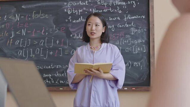 Tilt up shot of young Asian female student reading her notes to group mates while standing in front of chalkboard during programming class in college