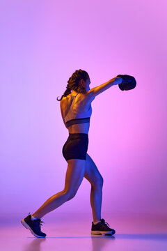 Striking image of female, professional sportsman, boxer showcasing her prowess in boxing gear against gradient studio background in neon filter.