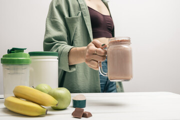 Young woman in jeans and shirt holding glass jar of protein drink cocktail, milkshake or smoothie above white wooden table with measuring spoon of protein powder, chocolate pieces, bananas and apples