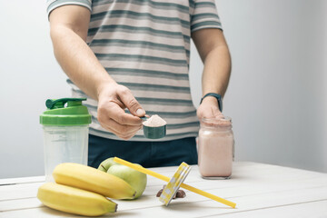 Man holding measuring spoon with protein powder, glass jar of protein drink cocktail, milkshake or smoothie above white wooden table with chocolate pieces, bananas, apples