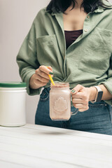 Young woman in jeans and shirt holding glass jar of protein drink cocktail, milkshake or smoothie...