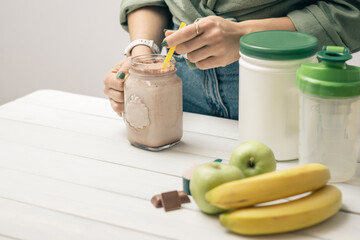 Young woman in jeans and shirt holding glass jar of protein drink cocktail, milkshake or smoothie above white wooden table with measuring spoon of protein powder, chocolate pieces, bananas and apples