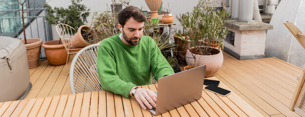 freelancer in earphone using laptop and working on rooftop terrace of house, banner